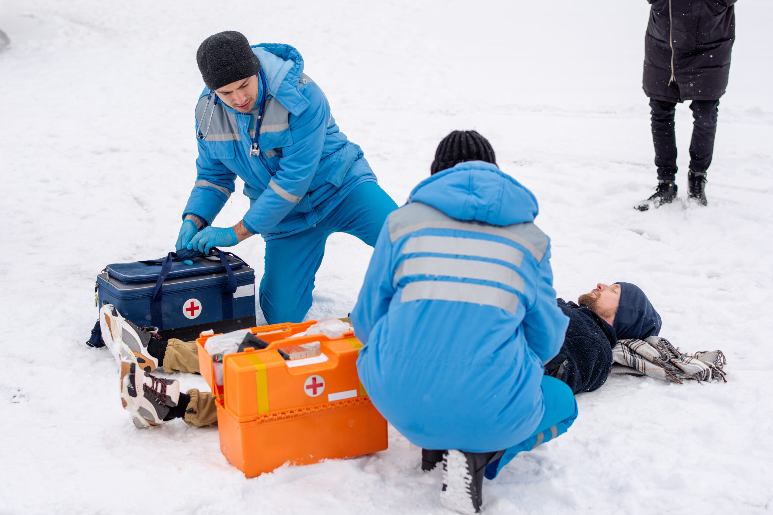 Two paramedics in uniform giving first aid to sick unconscious man lying on snow